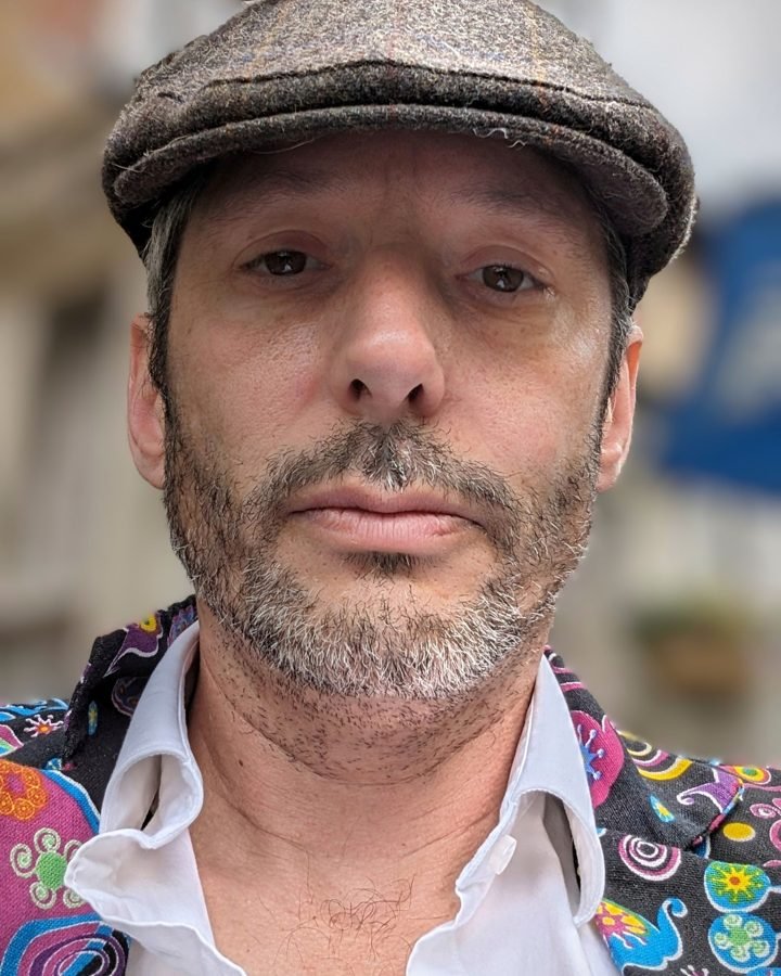 Adam Palk, wearing a flat cap and a brightly patterned waistcoat over a white shirt, looking straight at the camera